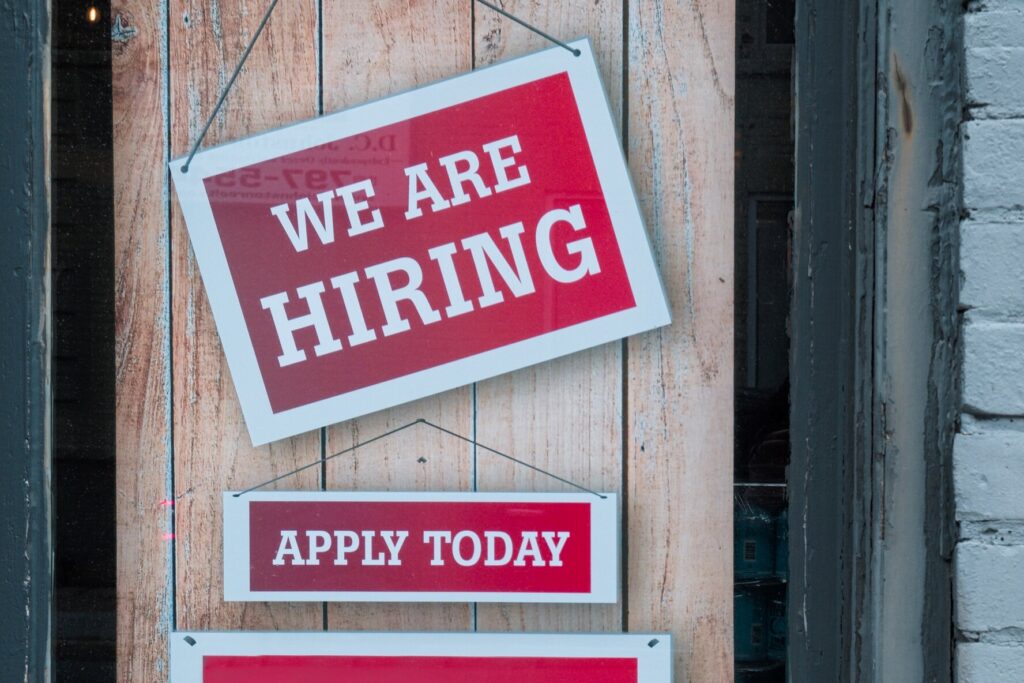 A wooden door with red and white "we are hiring" and "apply today" signs