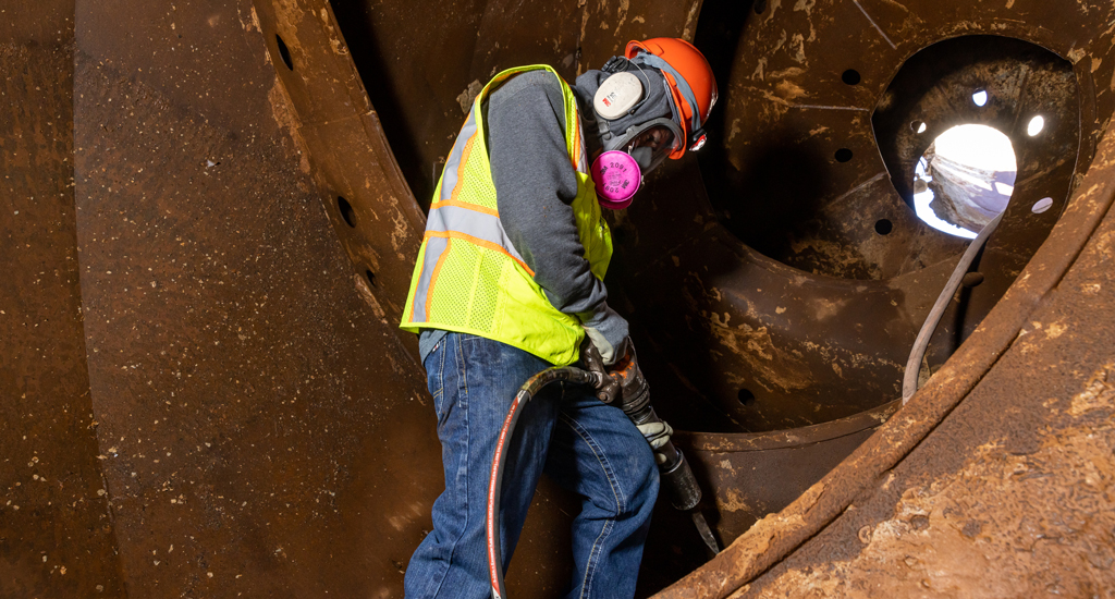 Man wearing PPE and holding a jackhammer, chipping concrete inside a ready mix concrete drum