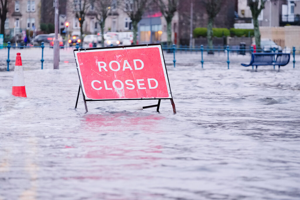 Red road closed sign in a flooded roadway with cars and buildings behind it