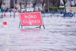 Red road closed sign in a flooded roadway with cars and buildings behind it