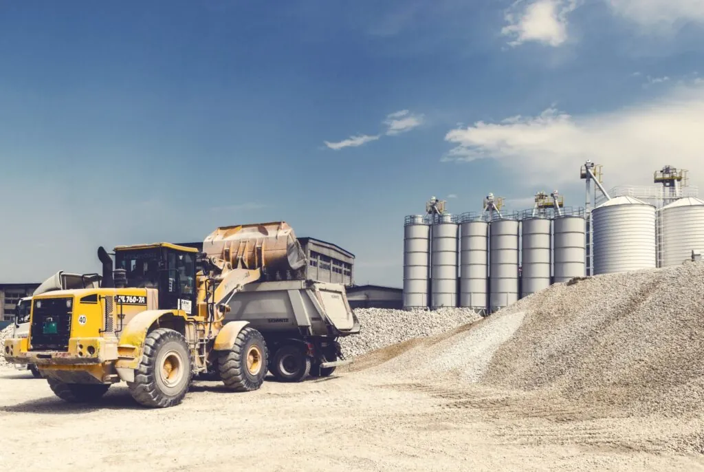 Vehicles hauling and moving gravel in front of cement storage silos at concrete plant